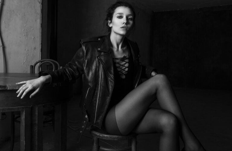 Black and white of young sensual woman in leather jacket sitting with crossed legs near table and looking at camera