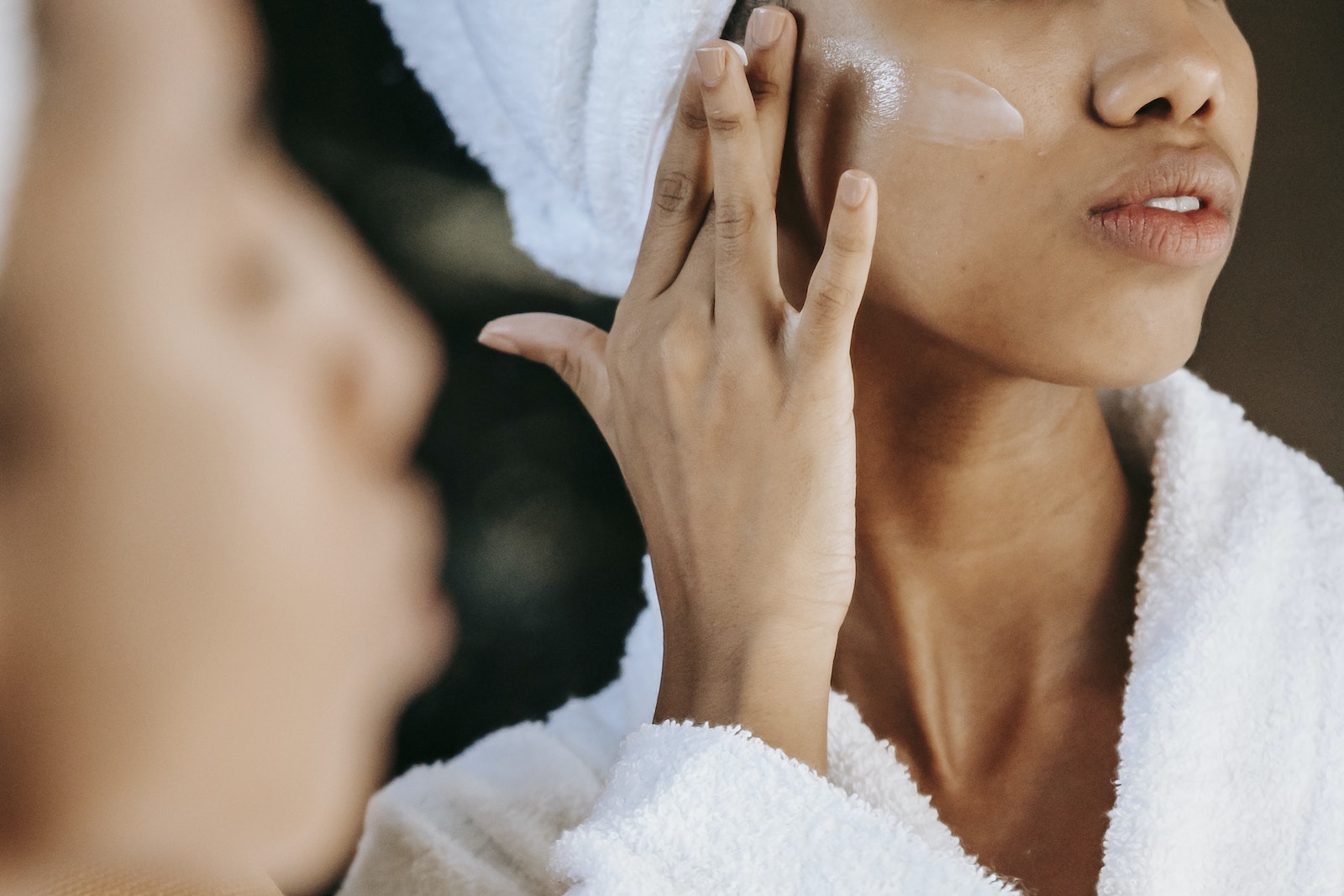 Crop unrecognizable young ethnic female in terry robe applying moisturizing cream on cheek while reflecting in mirror