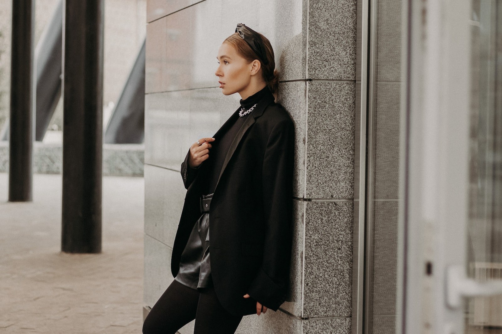 woman in black coat and black pants leaning on gray concrete wall