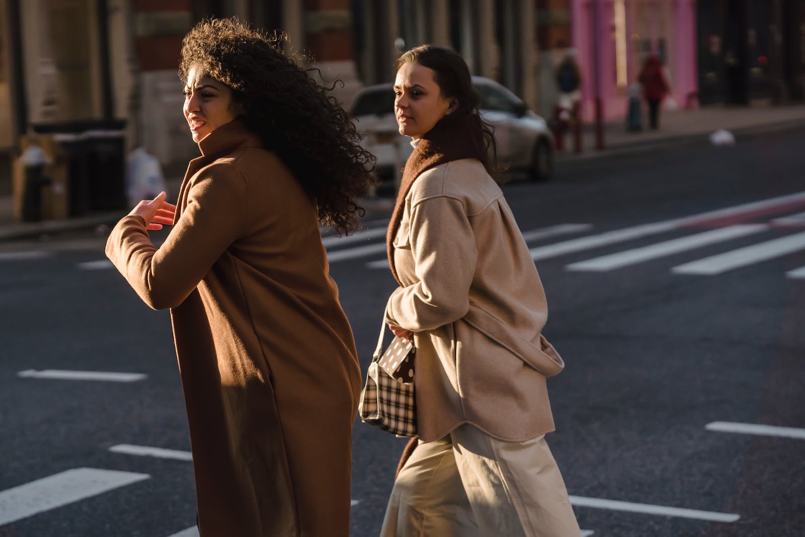 Stylish focused friends in fashionable coats walking across asphalt road with crosswalk in daytime