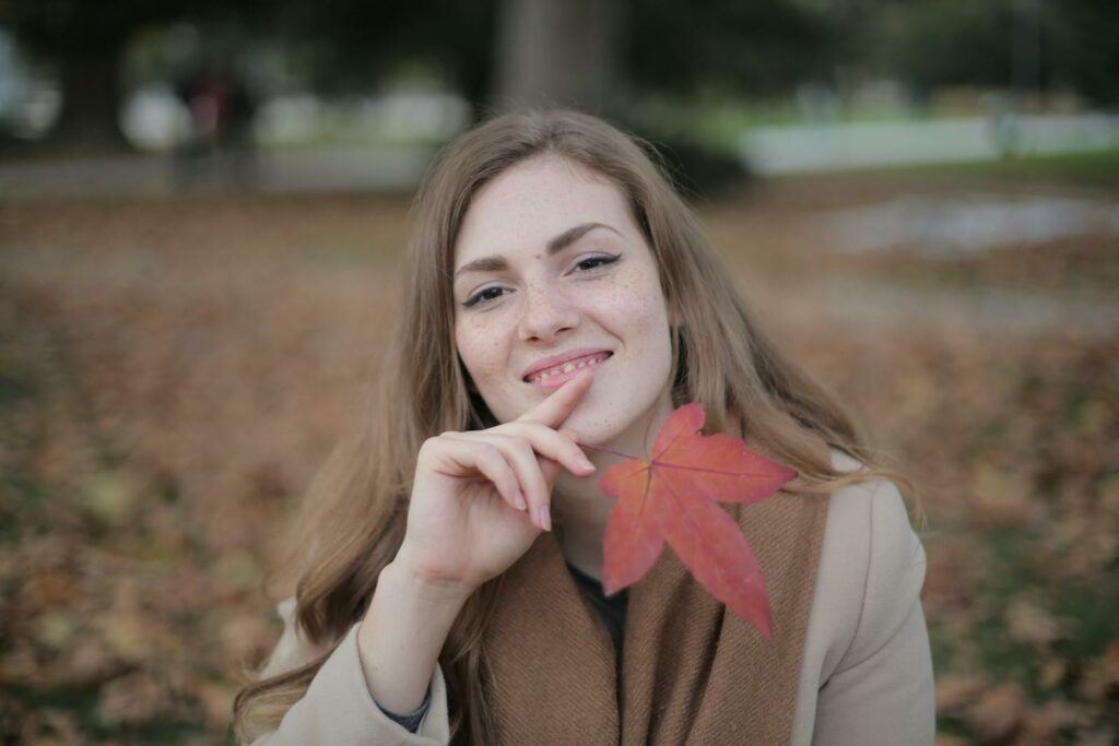 Attractive young lady in warm clothes and scarf with red maple leaf touching chin and smiling at camera against blurred environment in autumn park