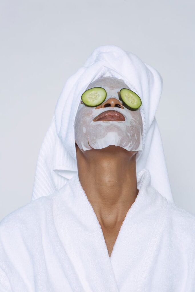 Unrecognizable black female in white bathrobe and towel on head and with sheet mask and cucumber slices on eyes standing head thrown back against white background