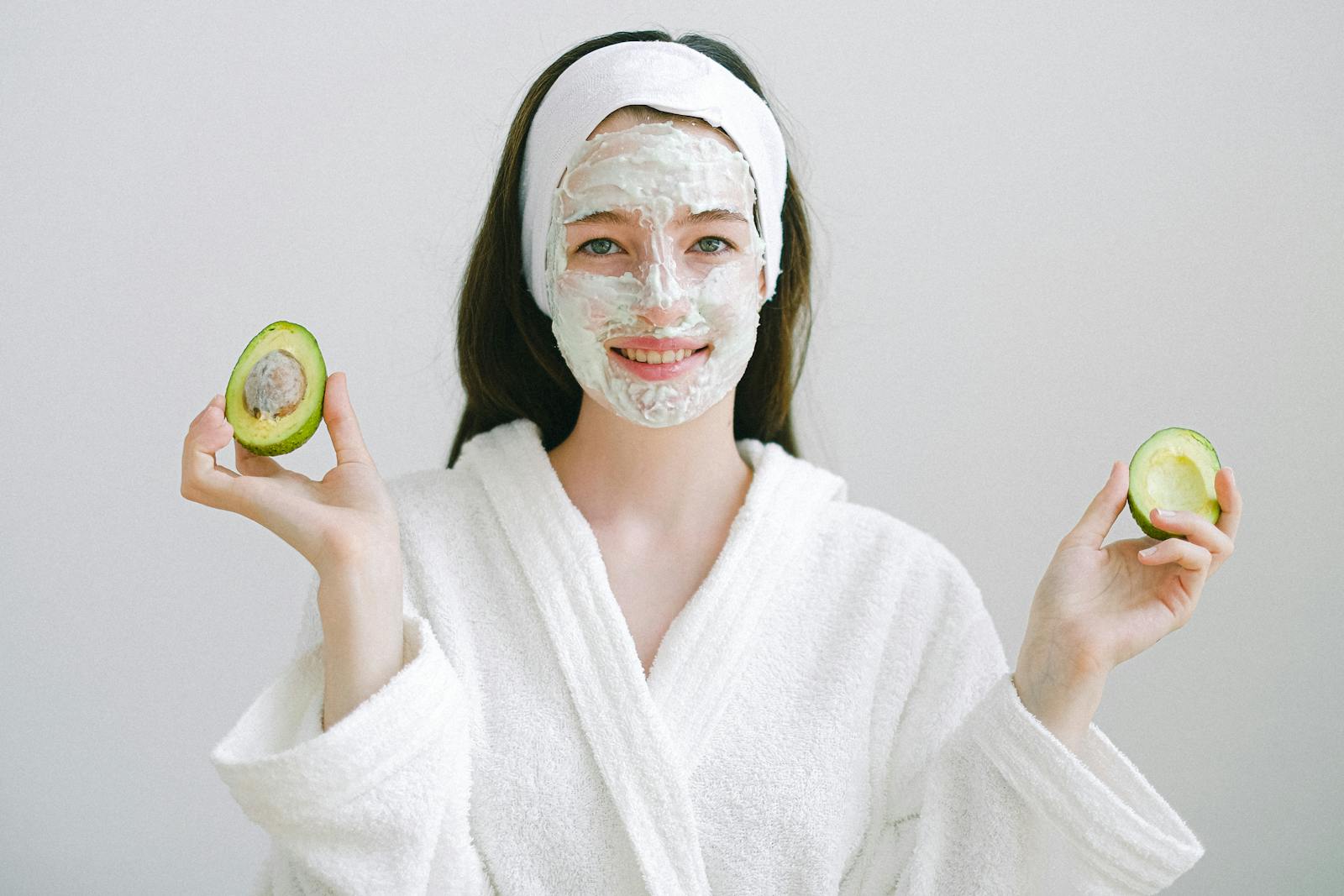 Cheerful female with facial mask wearing bathrobe and headband looking at camera while standing on white background with avocado in hands