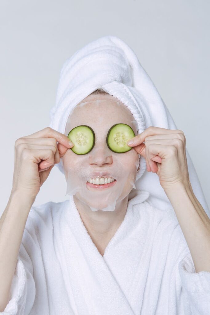 Positive female in towel turban and bathrobe applying cucumber slices on face with sheet mask for skincare treatment